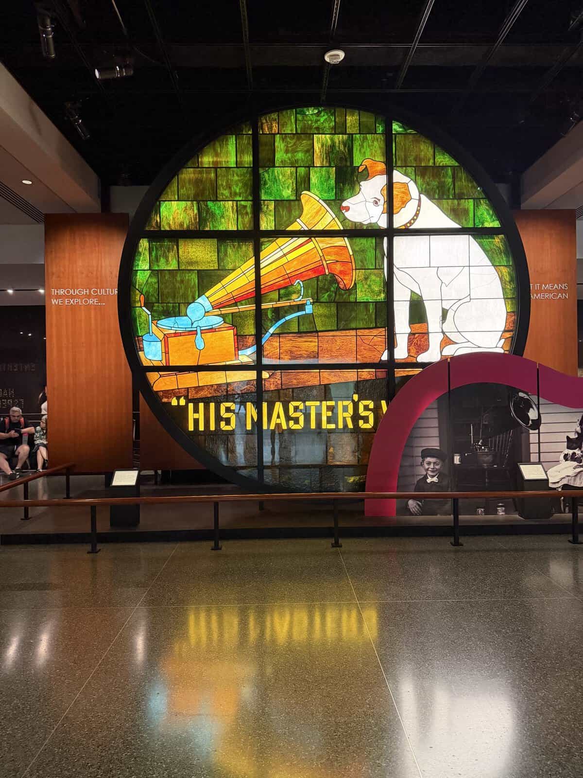 Large circular stained-glass display of a white dog listening to a gramophone with the text'HIS MASTER'S' at the bottom, museum exhibit.