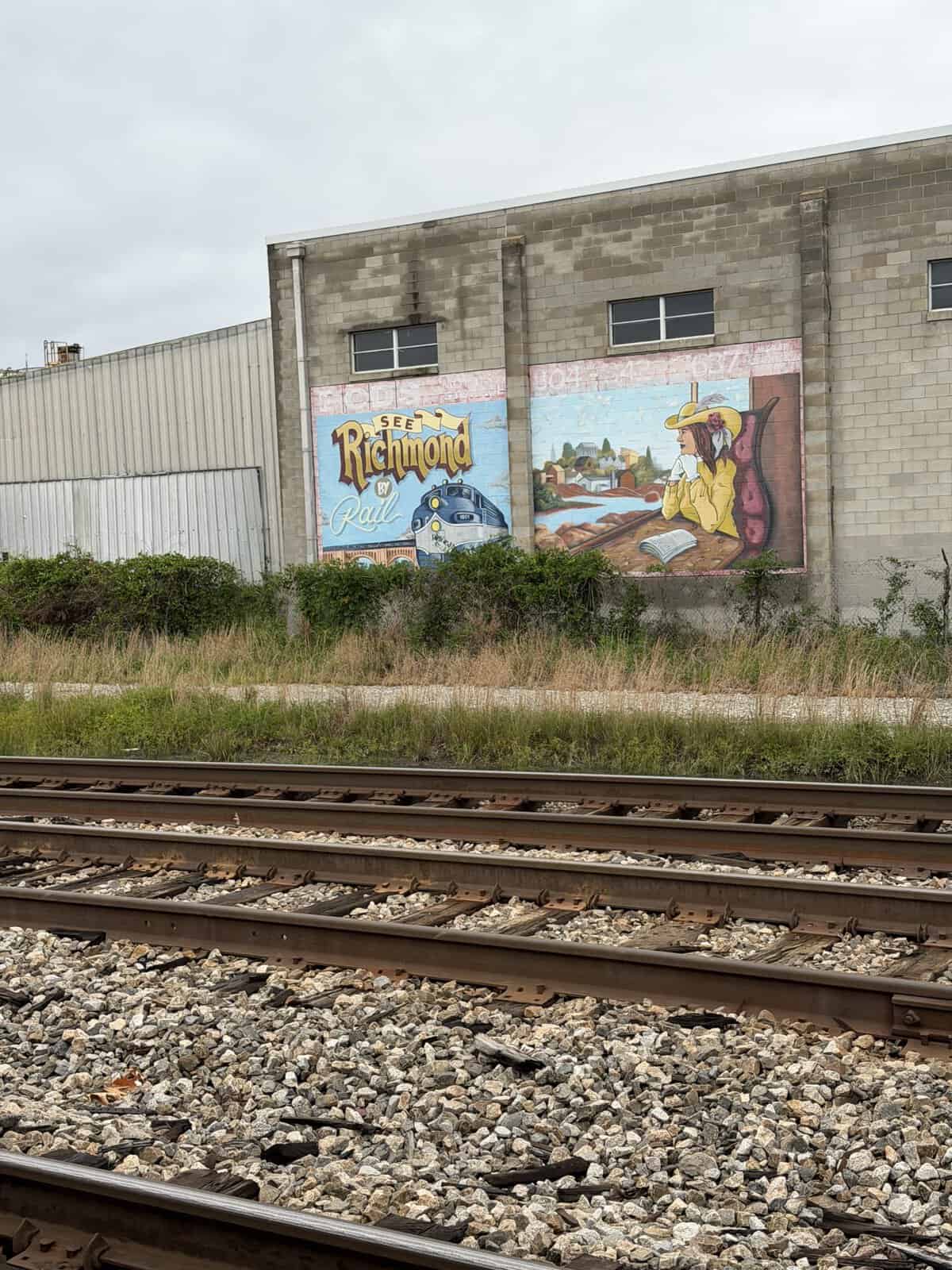 Brick industrial building with a large'Richmond by Rail' mural and a train illustration, beside railroad tracks.