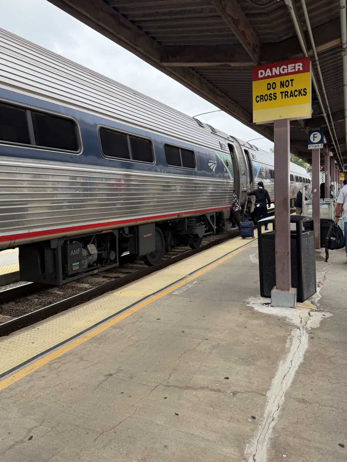Train at a station platform with passengers boarding a sleek silver passenger car beside a yellow danger sign that reads'Do Not Cross Tracks'