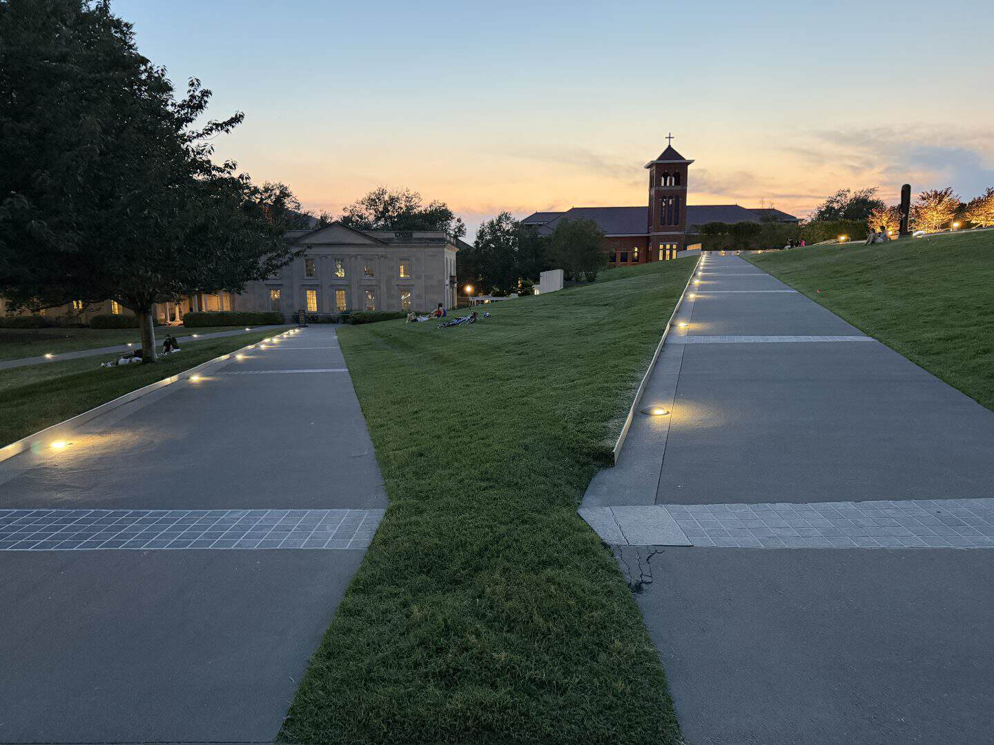 Richmond park walkway at dusk with a brick clock tower and a white/classical building, illuminated path lights along the concrete paths on a green lawn.