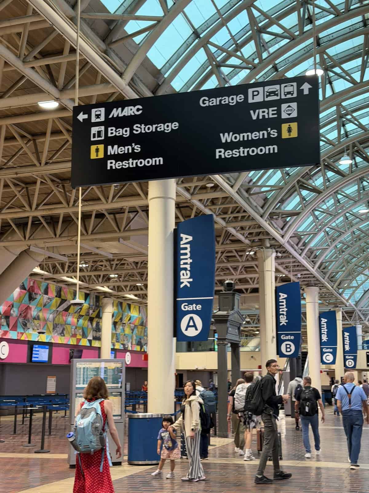 Large black directional sign in a busy train station listing MARC, Bag Storage, Men’s and Women’s Restrooms, Garage, and VRE; travelers walk underneath.