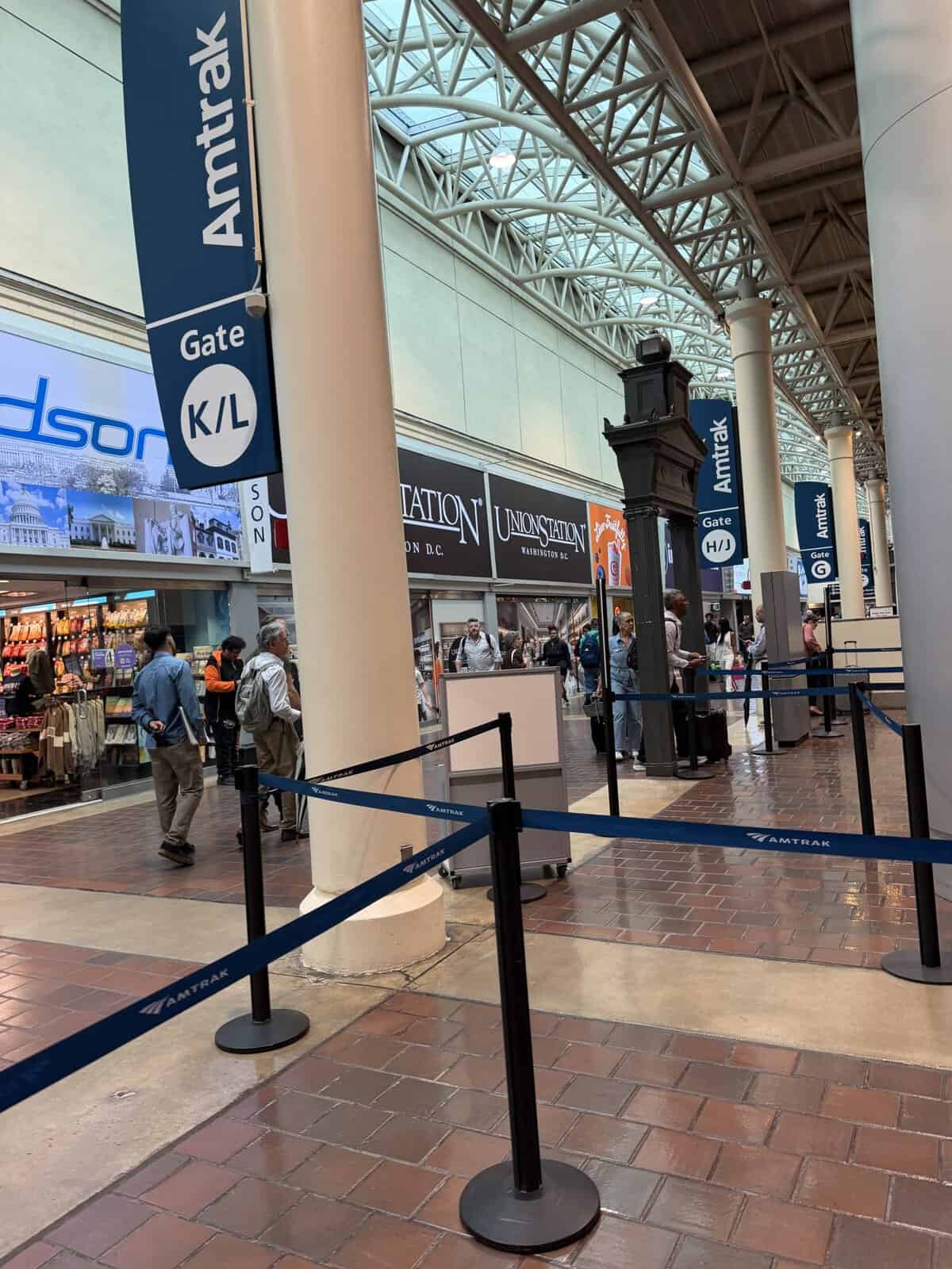 Busy train station concourse with Amtrak signs, blue gate barriers, and shoppers walking beneath a glass and steel roof.