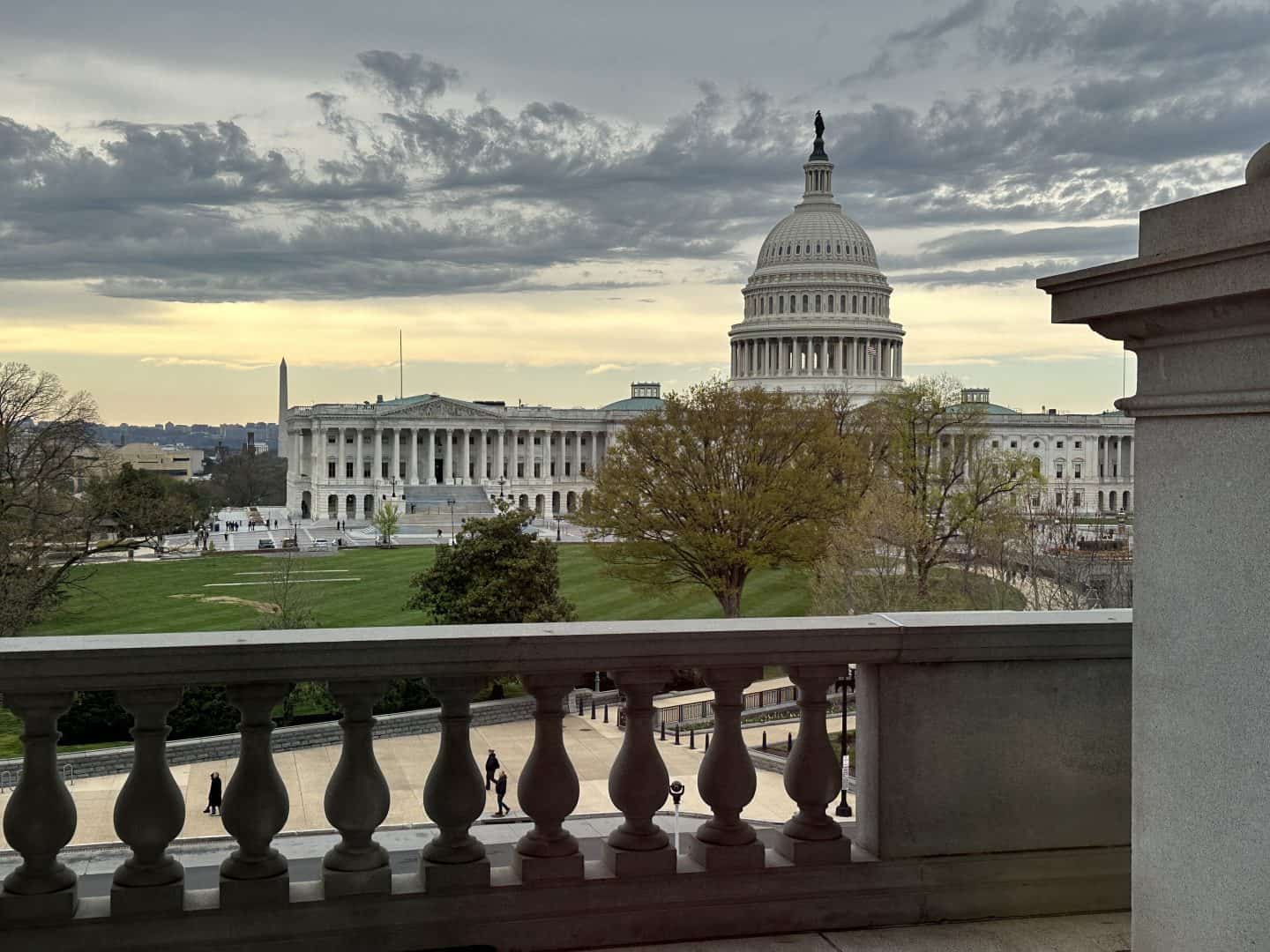 View of the U.S. Capitol with its dome, seen across a green lawn and trees from a balcony railing under a cloudy sky.